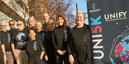 A group of lab professionals in UNI5K t-shirts standing next to a big sign that says "UNI5K - Unify for something greater."